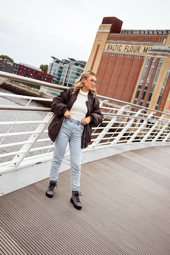 An image of a girl standing on a bridge wearing a pair of grey biker boots, blue jeans, white top and a brown bomber jacket