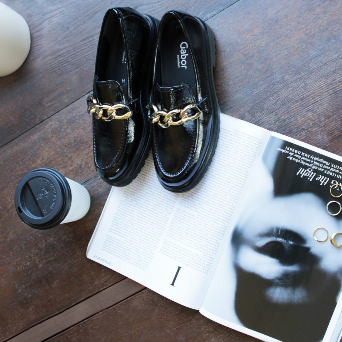 Loafers in black with a gold knot finish are pictured with an open magazine and coffee cup.