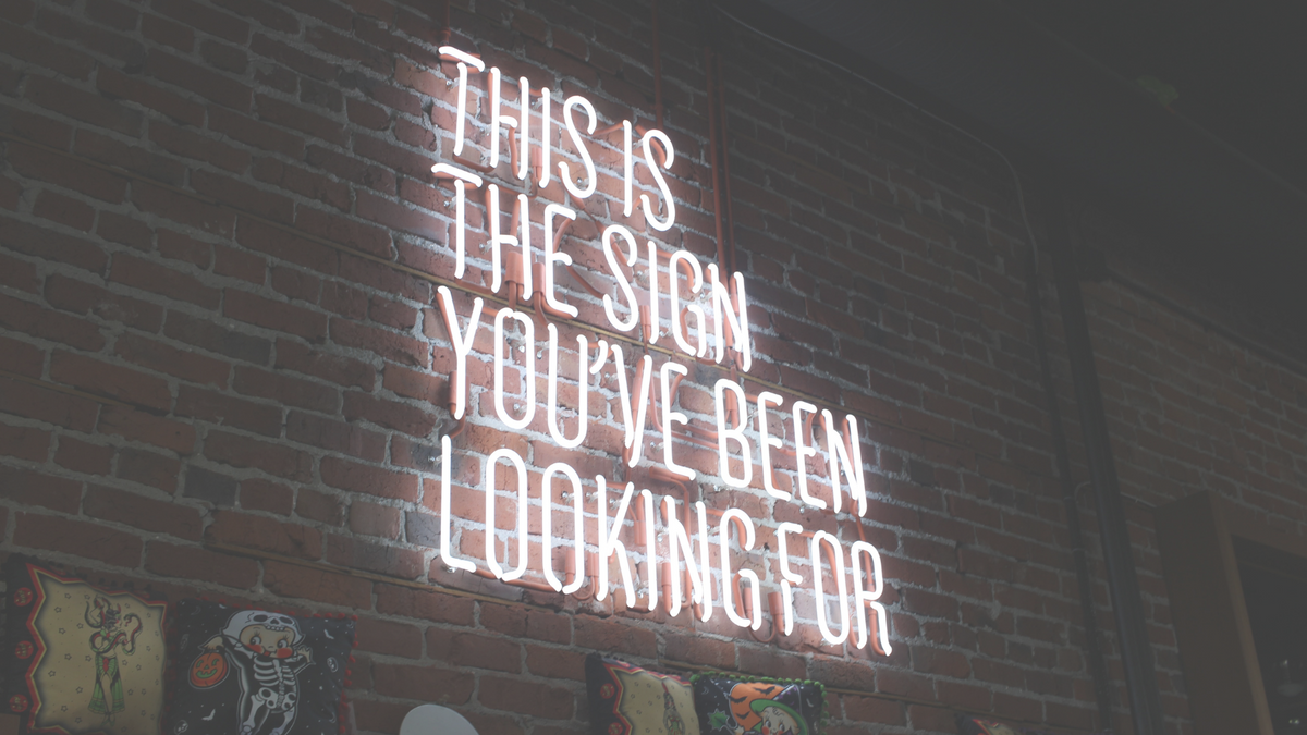 An image of a sign that reads "This is the sign you have been looking for" in a red neon colour is set against a dark wall.