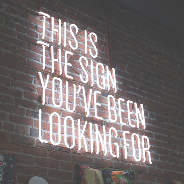 An image of a sign that reads "This is the sign you have been looking for" in a red neon colour is set against a dark wall.