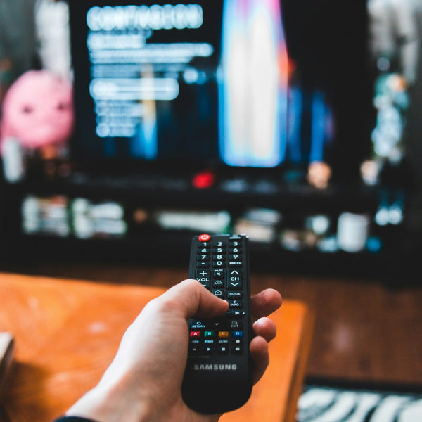 A hand holding a remote is in focus, and is pointing to a TV open on Netflix ready to watch a feel-good film