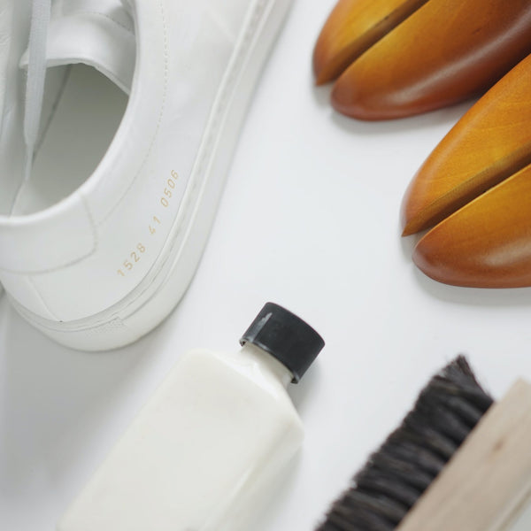 A heel of a pair of white shoes are in shot, with a cleaning brush and bottle of liquid also on display.